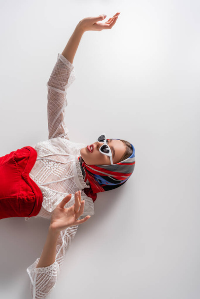 top view of stylish woman in headscarf and sunglasses lying with outstretched hand on white - Photo, Image