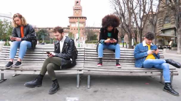 Młody mężczyzna kaukaski czyta książkę odróżniającą od innych wielonarodowych ludzi nieznajomi lub przyjaciele siedzący na ławce outdoor za pomocą smartfona ignorując uzależnionych od trendów społecznych - Materiał filmowy, wideo