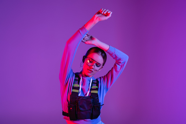 stylish woman in sunglasses with hands above head posing on purple  - Foto, afbeelding