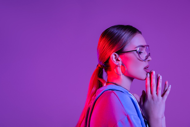 vista lateral de la joven mujer con estilo en gafas de sol y las manos de oración aisladas en púrpura  - Foto, imagen