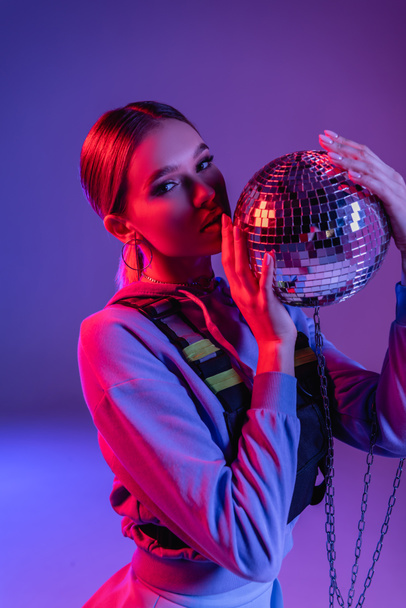 young and stylish woman holding shiny disco ball on purple - Foto, imagen