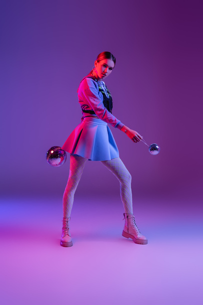 full length of fashionable woman in skirt holding disco balls on purple - Foto, immagini