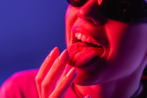 cropped view of woman in sunglasses putting paper heart on tongue isolated on purple - Фото, зображення