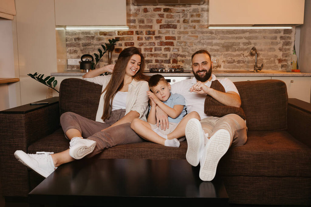 Papa, Sohn und Mama schauen auf dem Sofa in der Wohnung fern. Die Familie genießt einen fröhlichen Abend zu Hause. Eine junge Mutter mit langen Haaren lächelt. - Foto, Bild