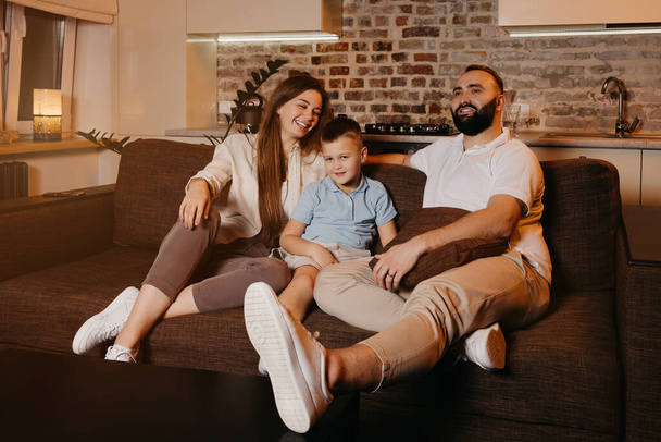 Папа с бородой, сын и молодая мама с длинными волосами смотрят телевизор и улыбаются на диване. Семья наслаждается счастливым вечером дома. Мать смеется. - Фото, изображение