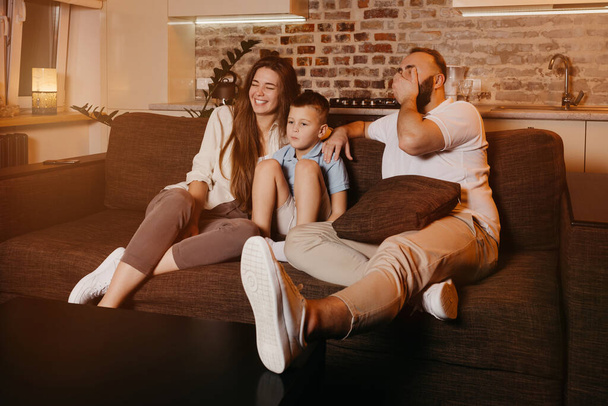 Táta s vousy, syn a mladá máma s dlouhými vlasy se dívají na televizi na gauči. Rodina si užívá šťastný večer doma. Otec se směje a zakrývá si tvář rukou.. - Fotografie, Obrázek