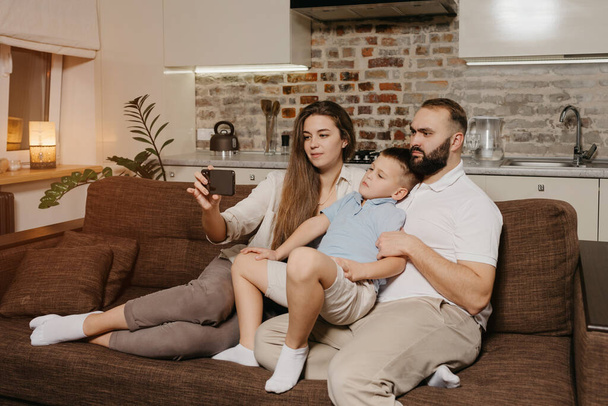 A dad, a son, and a young mother are watching a video on a smartphone on the sofa. A mom is demonstrating to her family news on the screen of the cellphone in the evening at home. - Photo, Image