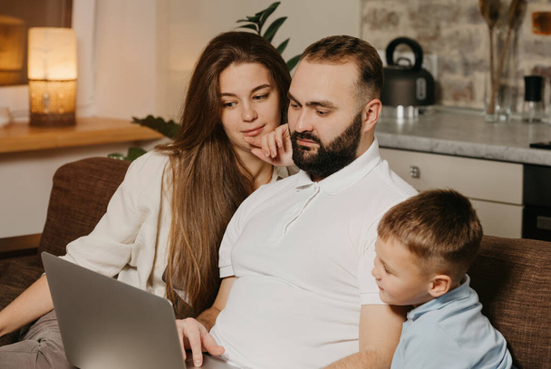 A close up photo of father with a beard who is showing his achievements at work on a laptop to his son and pensive wife at home. Dad is working remotely on a computer between relatives in the evening - Photo, Image