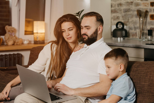 A father with a beard is talking about his achievements at work to his son and wife at home. A young family on the sofa in the evening. Dad is working remotely on a laptop between relatives - Photo, Image