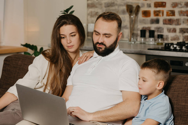 A father with a beard is talking about his achievements at work to a son and a wife at home. A family on the sofa in the evening. Dad is working remotely on a computer between relatives - Foto, immagini