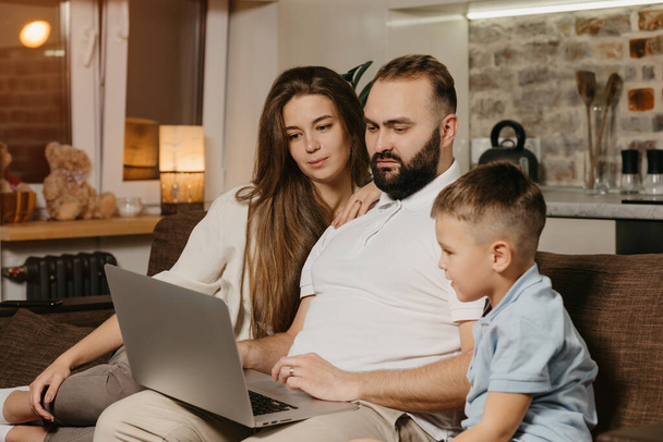 Otec s plnovousem pracuje na dálku na laptopu, zatímco jeho syn a žena na něj zírají. Večer je rodina na gauči. Táta pracuje online na počítači mezi příbuznými doma - Fotografie, Obrázek