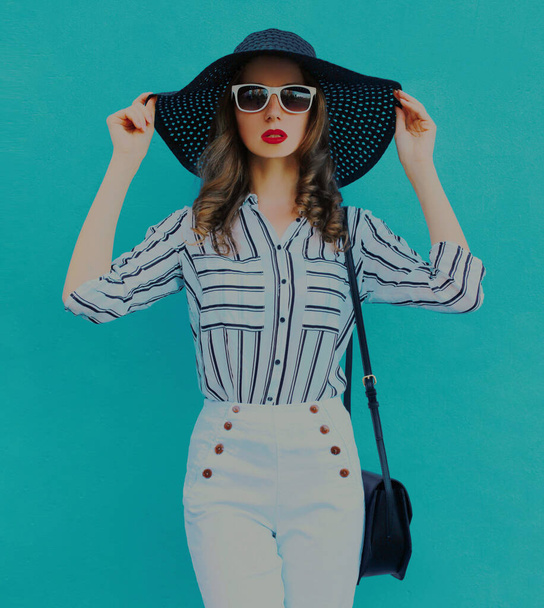 Portrait of beautiful young woman wearing a black round summer hat, white striped shirt on a blue background - Фото, зображення