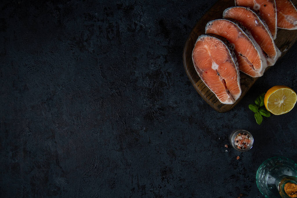Dark textured background with free space, with the edge ingredients: salmon steaks on a board, salt, pepper, oil and lemon. - Fotografie, Obrázek
