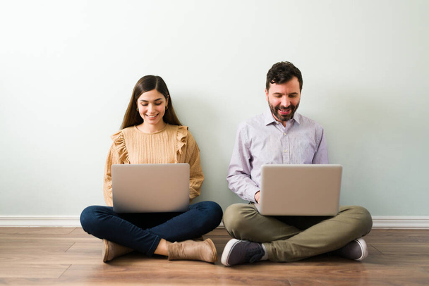 Atractiva pareja latina con ordenadores portátiles sentados juntos en el suelo y buscando un socio en una línea citas sitio web de amor  - Foto, imagen