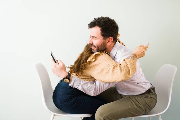 Hombre guapo de unos 30 años y mujer joven sentada y abrazada mientras sostiene un teléfono inteligente. Pareja usando una aplicación de citas en línea - Foto, imagen