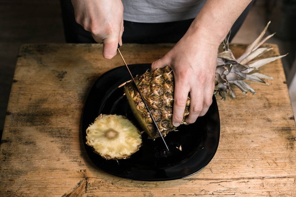 Man cuts pineapple with a knife on a wooden table - Valokuva, kuva