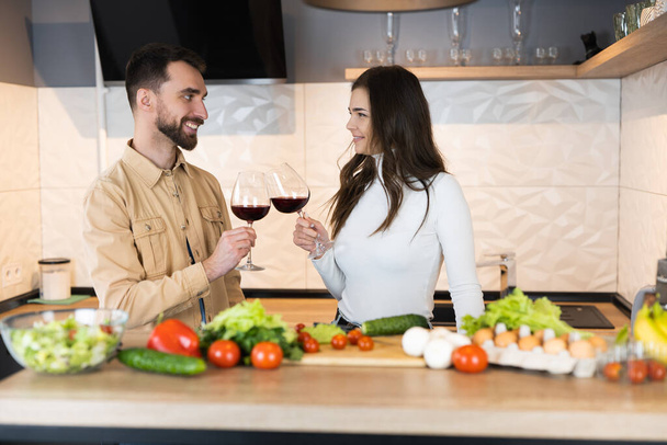 Bonito casal vegetariano estão cozinhando e bebendo vinho tinto juntos em uma data na cozinha doméstica. Também eles estão usando apenas produtos frescos e saudáveis - Foto, Imagem