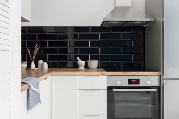 Small and stylish kitchen with white furniture, wooden countertop, black brick style tiles and silver oven, kitchen hood and fridge - Fotoğraf, Görsel
