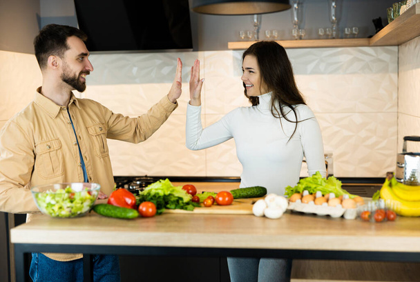 Χαριτωμένο χορτοφάγος ζευγάρι είναι hiving πέντε ο ένας τον άλλον, ενώ το μαγείρεμα μαζί και φαίνεται τόσο χαρούμενος και υγιής. Ευτυχισμένοι vegan άνθρωποι μαγειρεύουν σαλάτα χρησιμοποιώντας μόνο τη διατροφή και υγιεινά τρόφιμα. - Φωτογραφία, εικόνα