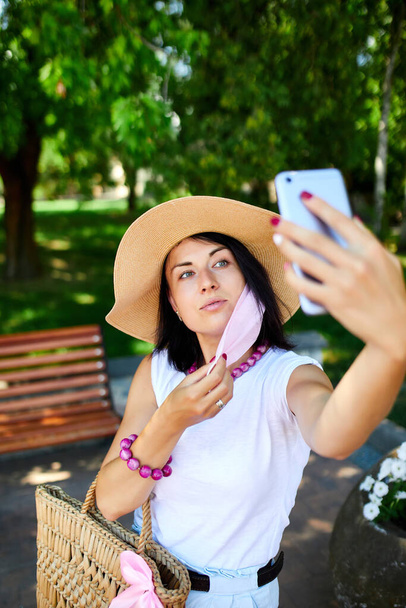Γυναίκα σε ροζ ιατρική μάσκα στο πάρκο λάβει selfie από το κινητό τηλέφωνο, Γυναίκα με αναπνευστική προστασία είναι σε εξωτερικούς χώρους, ενώ μιλάμε και να έχουν βίντεο chat με κάμερα στο smartphone - Φωτογραφία, εικόνα