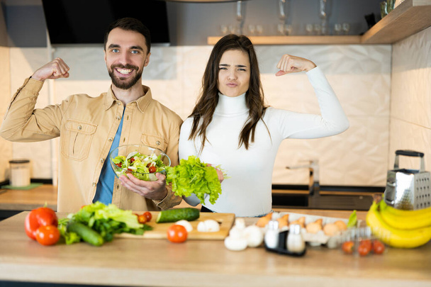 Cute vegetarian couple is showing how healthy and strong they are. Happy vegan people are cooking salad using only nutrition and healthy food - Photo, Image