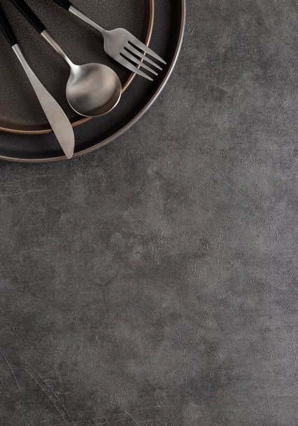 Fork, spoon and knife on dark round ceramic plate on dark gray concrete background with copy space. Top down view, flat lay, place for text, template - Foto, afbeelding