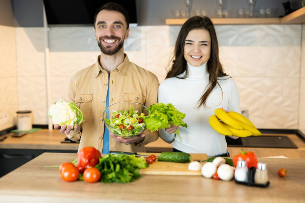 Genç ve güzel vejetaryen çift mutfakta yemek pişirirken gülümsüyor. Ayrıca sadece besin ve taze yiyecek kullandıkları için mutlu ve sağlıklı görünüyorlar. - Fotoğraf, Görsel