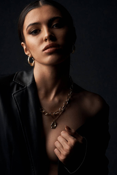 portrait photo of a dark-eyed beautiful girl with black hair, professional make-up, dressed in a black jacket, she has gold jewelry on her arm, ears and neck - Photo, Image