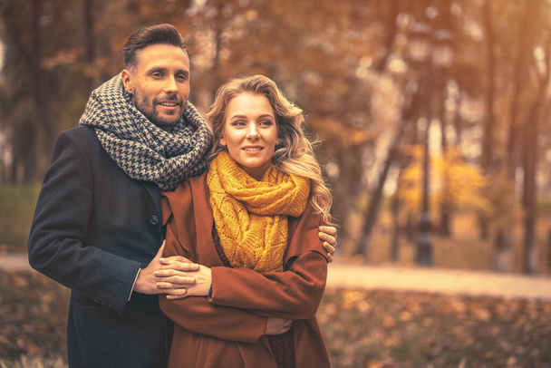 Couple in love man and a woman hugged from behind smile looking at their kids in the autumn park. Outdoor shot of a young couple in love having great time. Autumn toned image - Zdjęcie, obraz