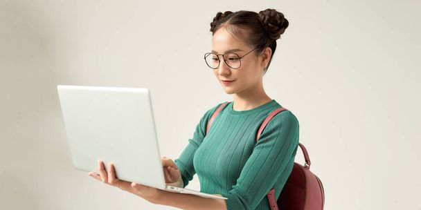 Portrait of a pretty cheerful casual asian girl carrying backpack standing isolated over white background, holding laptop computer - Fotó, kép