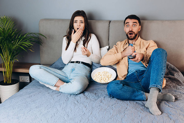Omg what is it. Sweet couple has quarantine free time watch movie impressed surprised unexpected thriller film ending with pop corn box sit sofa in house indoors. Lovely shocked couple watching tv - Fotografie, Obrázek