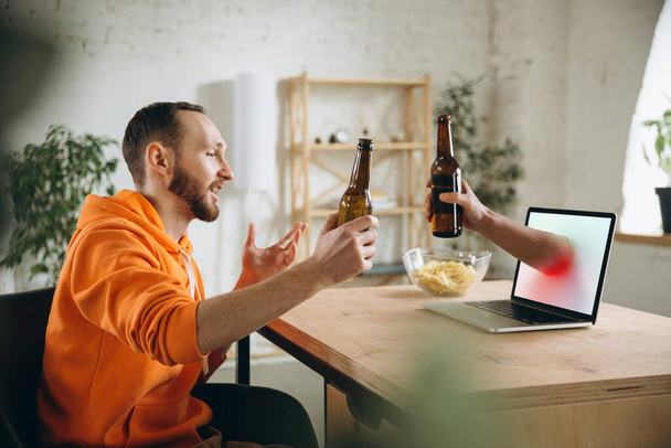 Young man drinking beer during meeting friends on virtual video call. Distance online meeting, chat together on laptop at home. - Valokuva, kuva