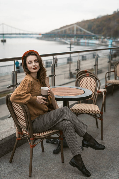 Sitting sideways turned Parisian young woman in restaurant terrace. Portrait of stylish young woman wearing autumn coat and red beret outdoors - Фото, изображение