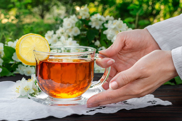 Las manos femeninas sirven una taza de vidrio con té, limón, servilleta bordada, ramas de jazmín en una mesa de madera. Al aire libre, picnic, almuerzo. - Foto, Imagen