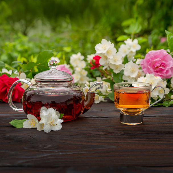 Egy párolt teáskanna és egy elegáns üveg csésze között virágzó rózsa és jázmin bokrok. Kint, piknik, villásreggeli. Zöld levelek háttér elmosódott. - Fotó, kép