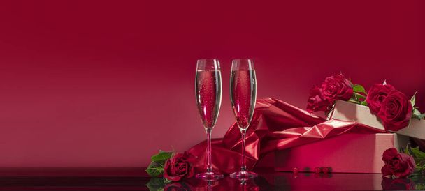 Luxurious glasses with sparkling wine open box with glitter brocade surrounded by selected roses on a mirror surface on a red background. Valentine's day or romantic evening invitation - Photo, Image
