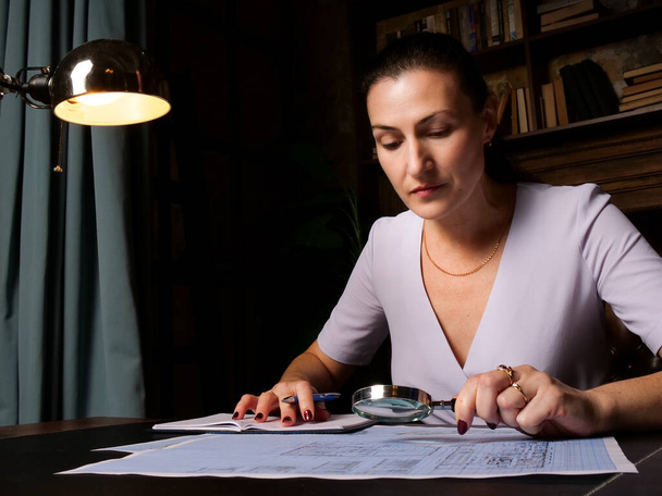 Konzentrierte Geschäftsfrau sitzt am Schreibtisch und prüft mit der Lupe eine Konstruktionszeichnung. Wirtschaftsprüfer verwenden Lupe für Papierkrieg Investigatio - Foto, Bild
