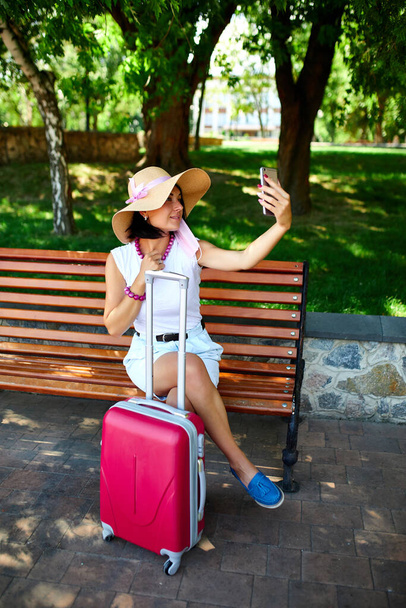 Blogger woman in straw hat and removed mask holds on her head, in the park outdoor with a malitcase, take selfie, life during coronavirus pandemic, opening air travel, travel concept. - Foto, Imagen