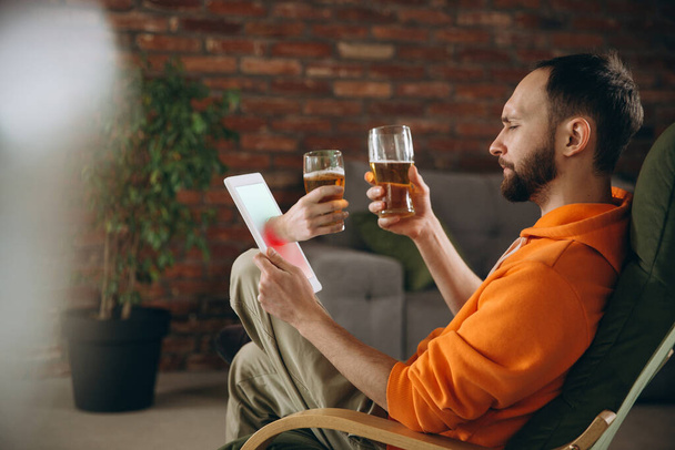 Молодой человек пьет пиво во время встречи с друзьями на виртуальном видео-вызове. Дистанционная онлайн-встреча, общение на планшете дома. - Фото, изображение