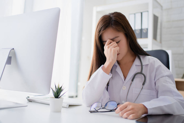 Asiatique femme médecin assis au bureau avec les yeux fermés et ayant mal à la tête après une dure journée de travail. - Photo, image