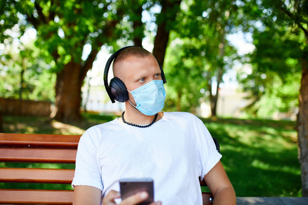 Чоловік слухає музику з навушниками, використовуючи смартфон у захисній масці на відкритому повітрі в парку, новий спосіб життя, карантин, коронавірус
. - Фото, зображення