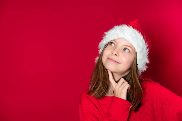 pretty young school girl with Santa Claus hat and red sweater in front of red background - Fotografie, Obrázek