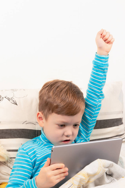 kind jongen in een blauwe trui thuis in bed speelt op de tablet op de achtergrond van een witte muur - Foto, afbeelding