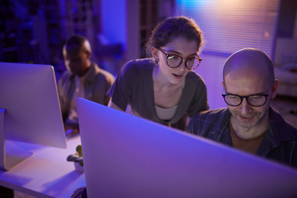 Vysoký úhel záběru moderní mladé ženy a jejího dospělého mužského kolegy sledující něco na obrazovce počítače v práci večer - Fotografie, Obrázek