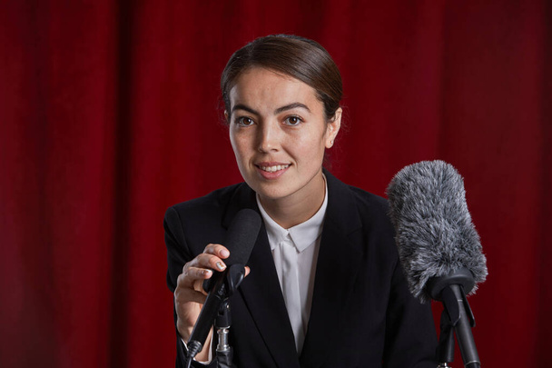 Portret młodej kobiety mówiącej do mikrofonu na scenie stojącej na podium o czerwonej kurtynie - Zdjęcie, obraz