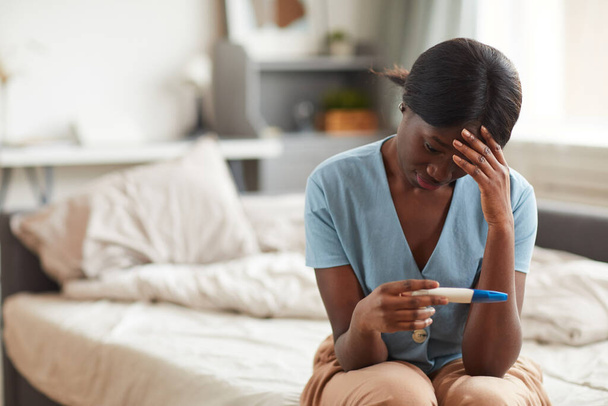 Portrait of worried African-American woman looking at pregnancy test while sitting on bed at home, copy space - Photo, Image