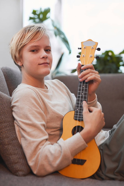 Retrato vertical del adolescente rubio sosteniendo el ukelele y mirando a la cámara mientras se relaja en el sofá en casa - Foto, Imagen