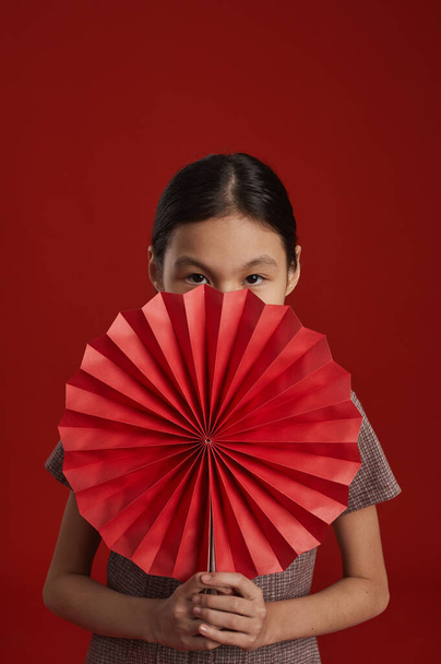 irreconocible asiático chica de pie sobre rojo pared fondo holding tradicional chino papel ventilador buscando cámara - Foto, imagen