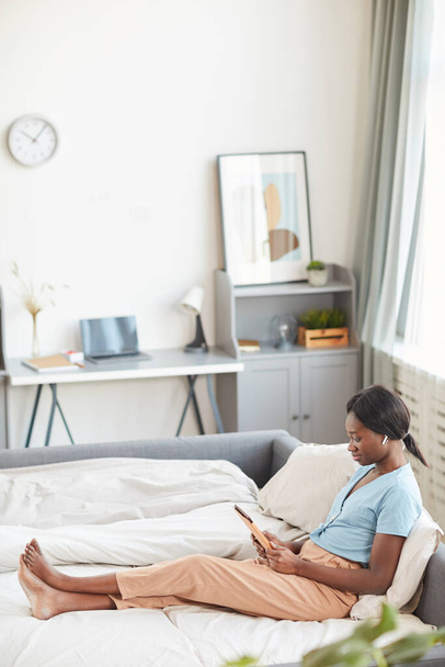 Широкоугольный вертикальный портрет молодой афроамериканки, лежащей утром на кровати и слушающей музыку через беспроводные наушники, копировальное пространство - Фото, изображение