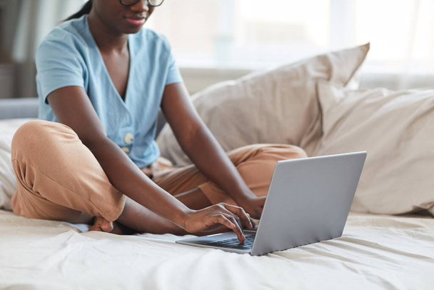 Καλλιεργημένο πορτρέτο της νεαρής Αφροαμερικανής γυναίκας που χρησιμοποιεί φορητό υπολογιστή, ενώ κάθεται στο κρεβάτι στο σπίτι, αντιγραφή χώρου - Φωτογραφία, εικόνα
